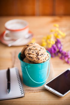 Chocolate chip cookies in small green bucket with smartphone, notebook and pen, and coffee cup at cafe in morning time