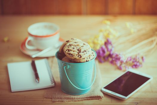 Chocolate chip cookies in small green bucket with smartphone, notebook and pen, and coffee cup at cafe in morning time with film filter effect
