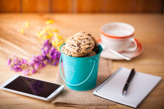 Chocolate chip cookies in small green bucket with smartphone, notebook and pen, and coffee cup at cafe in morning time