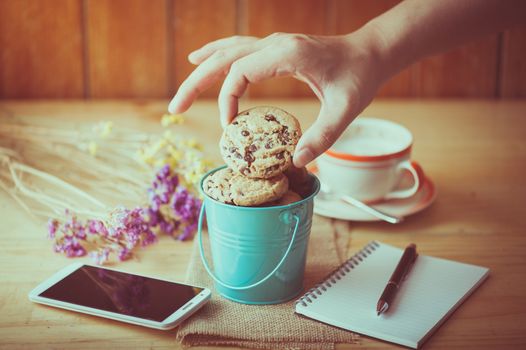 Male right hand taking chocolate chip cookie from small green bucket with smartphone, notebook and pen, and coffee cup at cafe in morning time with vintage filter effect