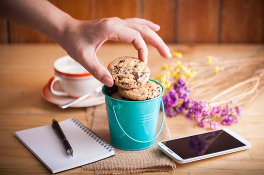 Male left hand taking chocolate chip cookie from small green bucket with smartphone, notebook and pen, and coffee cup at cafe in morning time