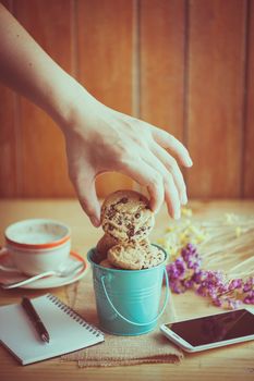 Male left hand taking chocolate chip cookie from small green bucket with smartphone, notebook and pen, and coffee cup at cafe in morning time with vintage filter effect