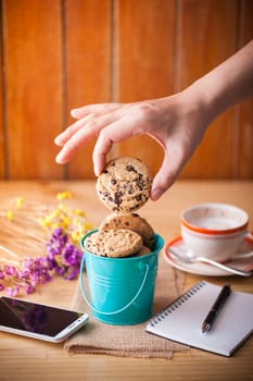 Male right hand taking chocolate chip cookie from small green bucket with smartphone, notebook and pen, and coffee cup at cafe in morning time