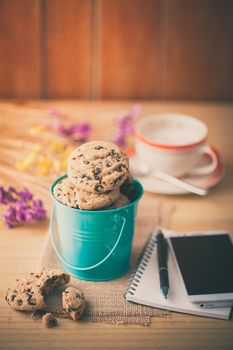 Chocolate chip cookies in small green bucket with smartphone, notebook and pen, and coffee cup at cafe in morning time with vintage filter effect