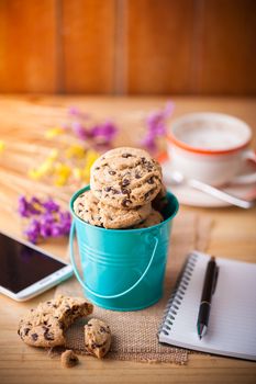 Chocolate chip cookies in small green bucket with smartphone, notebook and pen, and coffee cup at cafe in morning time