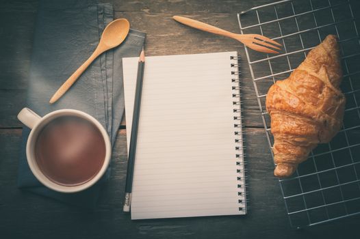 Butter croissant with a cup of hot chocolate and small notebook with blank area for text or message on wood table in morning time with film filter effect