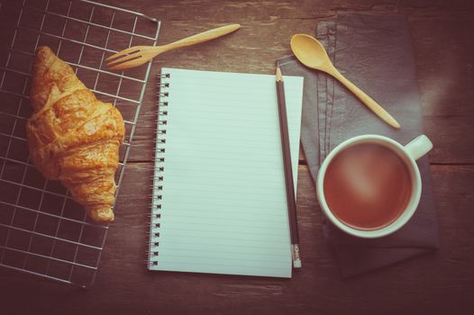 Butter croissant with a cup of hot chocolate and small notebook with blank area for text or message on wood table in morning time with film filter effect