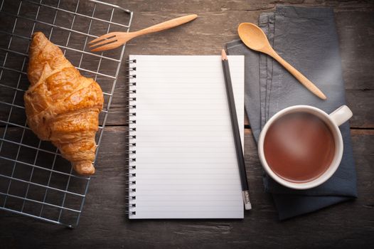 Butter croissant with a cup of hot chocolate and small notebook on wood table in morning scene with blank copyspace area for text or message
