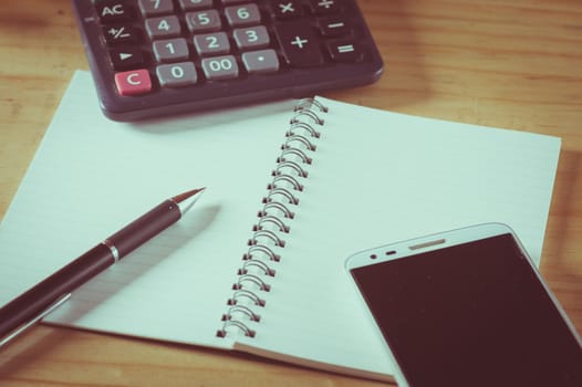 Opened notepad with blank area for text or message, pen, smart phone, and calculator on wood table with film filter effect