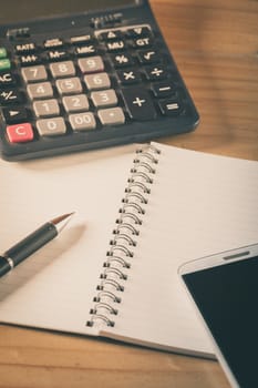 Opened notepad with blank area for text or message, pen, smart phone, and calculator on wood table with film filter effect
