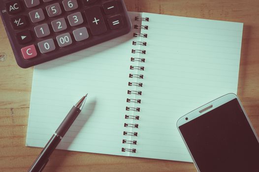 Opened notepad with blank area for text or message, pen, smart phone, and calculator on wood table with film filter effect