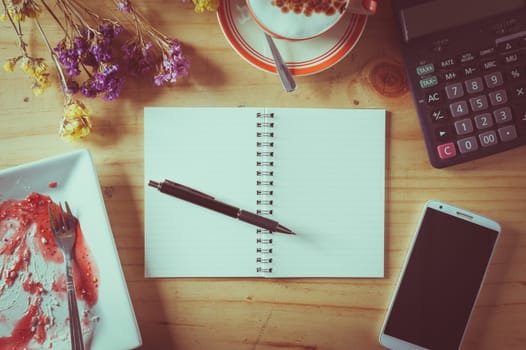 Opened notebook with blank area for text or message, pen, smart phone, dirty dessert dish, and calculator on wood table in afternoon time with film filter effect