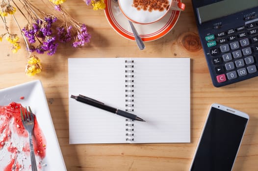 Opened notebook with blank area for text or message, pen, smart phone, dirty dessert dish, and calculator on wood table in afternoon time