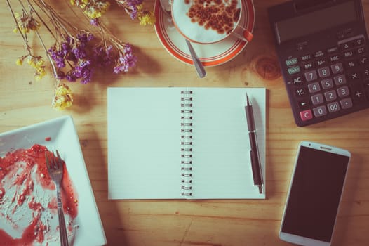 Opened notebook with blank area for text or message, pen, smart phone, dirty dessert dish, and calculator on wood table in afternoon time with film filter effect