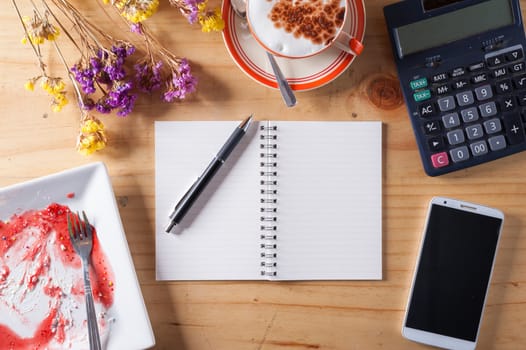 Opened notebook with blank area for text or message, pen, smart phone, dirty dessert dish, and calculator on wood table in afternoon time