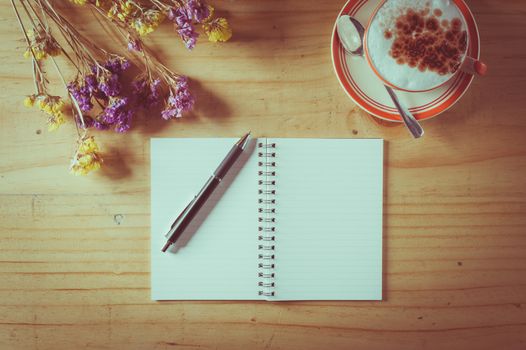 Opened notebook with blank area for text or message, pen and a cup of hot coffee on wood table in afternoon time with film filter effect