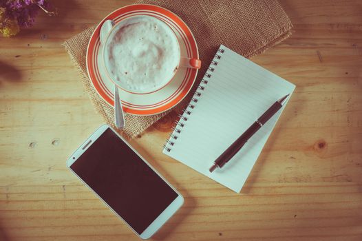 Opened notepad with blank area for text or message, pen, smart phone, and a cup of hot coffee on wood table in afternoon time with film filter effect