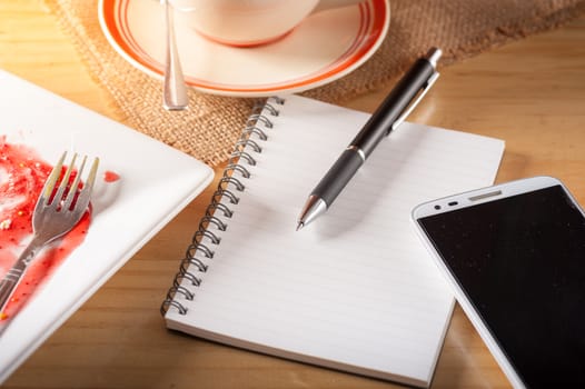 Opened notepad with blank area for text or message, pen, smart phone, and a cup of hot coffee on wood table in afternoon time