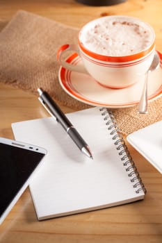 Opened notepad with blank area for text or message, pen, smart phone, and a cup of hot coffee on wood table in afternoon time