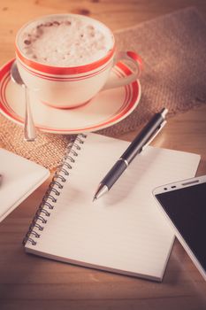 Opened notepad with blank area for text or message, pen, smart phone, and a cup of hot coffee on wood table in afternoon time with film filter effect
