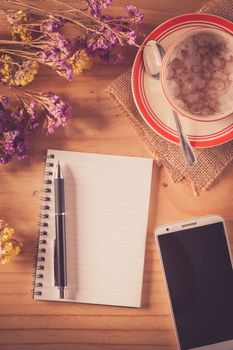 Opened notepad with blank area for text or message, pen, smart phone, and a cup of hot coffee on wood table in afternoon time with film filter effect