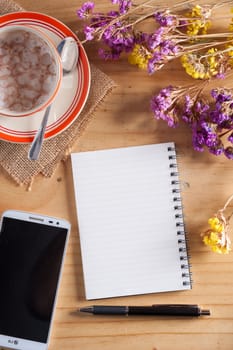 Opened notepad with blank area for text or message, pen, smart phone, and a cup of hot coffee on wood table in afternoon time