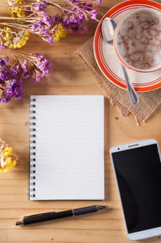 Opened notepad with blank area for text or message, pen, smart phone, and a cup of hot coffee on wood table in afternoon time