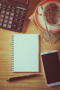 Opened notepad with blank area for text or message, pen, smart phone, and calculator on wood table in morning time with film filter effect
