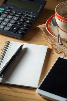 Opened notepad, pen, smart phone, and calculator on wood table in evening time with low key scene