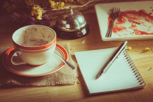 Opened notepad with blank area for text or message, pen, smart phone, and a cup of hot coffee on wood table in morning time with low key scene and film filter effect