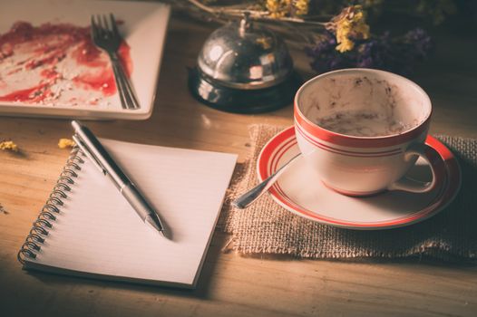 Opened notepad with blank area for text or message, pen, smart phone, and a cup of hot coffee on wood table in morning time with low key scene and film filter effect