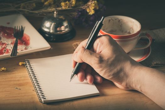Male right hand writing on opened notepad with blank area for text or message, pen, smart phone, and a cup of hot coffee on wood table in morning time with low key scene and film filter effect