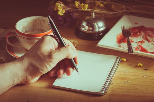 Male left hand writing on opened notepad with blank area for text or message, pen, smart phone, and a cup of hot coffee on wood table in morning time with low key scene and film filter effect