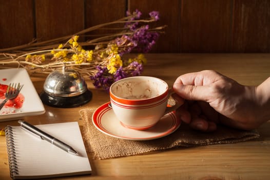 Male right hand holding a cup of coffee in cafe with notebook and pen on table in morning time with low key scene