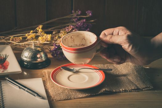 Male right hand holding a cup of coffee in cafe with notebook and pen on table in morning time with low key scene and film filter effect