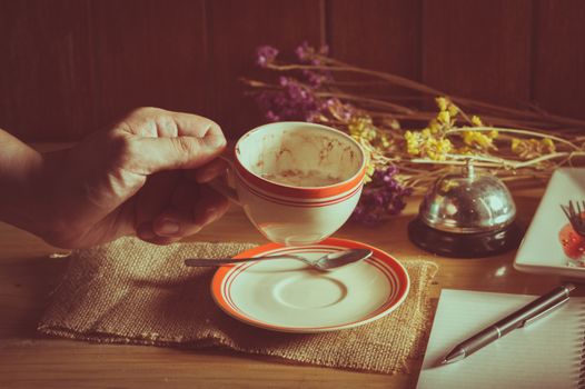 Male left hand holding a cup of coffee in cafe with notebook and pen on table in morning time with low key scene and film filter effect