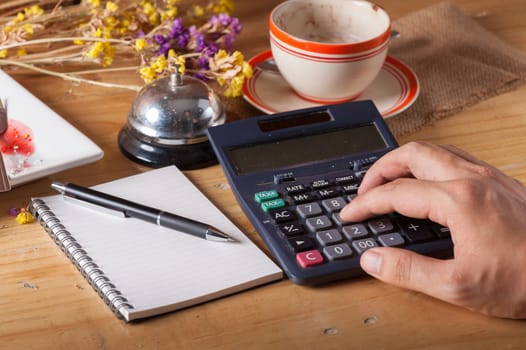 Male hand using calculator with opened notepad, pen, smart phone, and calculator on wood table in evening time with low key scene