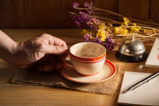 Male left hand holding a cup of coffee in cafe with notebook and pen on table in morning time with low key scene