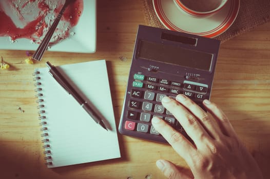 Male hand using calculator with opened notepad, pen, smart phone, and calculator on wood table in evening time with low key scene and film filter effect