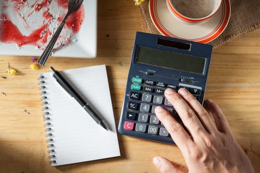 Male hand using calculator with opened notepad, pen, smart phone, and calculator on wood table in evening time with low key scene