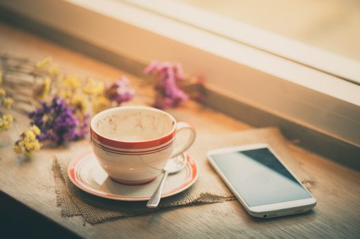 Coffee cup and smart phone on wood bar beside window at coffee shop in afternoon time with film filter effect