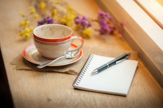 Coffee cup, opened notebook, and pen on wood bar beside window at coffee shop in afternoon time