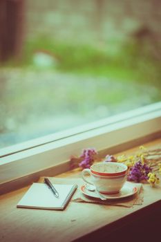Coffee cup, opened notebook, and pen on wood bar beside window at coffee shop in afternoon time
