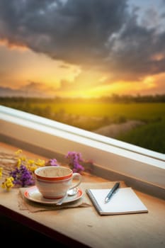 Coffee cup, opened notebook, and pen on wood bar beside window at coffee shop in morning time with nature view of sunrise