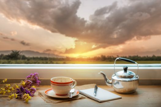 Coffee cup and opened notebook, and pen on wood bar beside window at coffee shop in morning time with nature view of sunrise