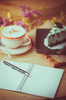 Opened notebook and pen, peppermint with chocolate cookie cheese cake, and hot cappuccino at cafe in afternoon time with film filter effect