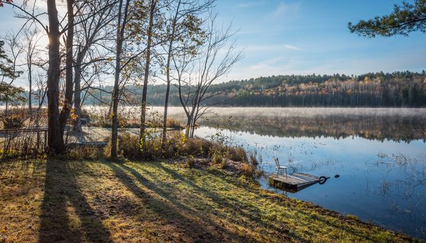 Morning sun rise revealing light fog.  Chair sits on a partially submerged dock beside a lake in Ontario, Canada.