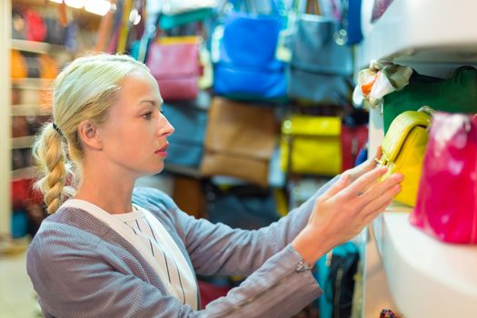 Beautiful casually dressed caucasian blond woman shopping for colorful new leather purse in fashion boutique.