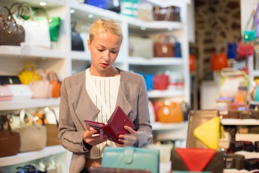 Beautiful casually dressed caucasian blond woman shopping for new colorful stylish leather wallet in fashion boutique.