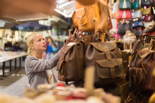 Beautiful casually dressed caucasian blond woman shopping for new leather bag on crafts market in Chania, Crete, Greece.